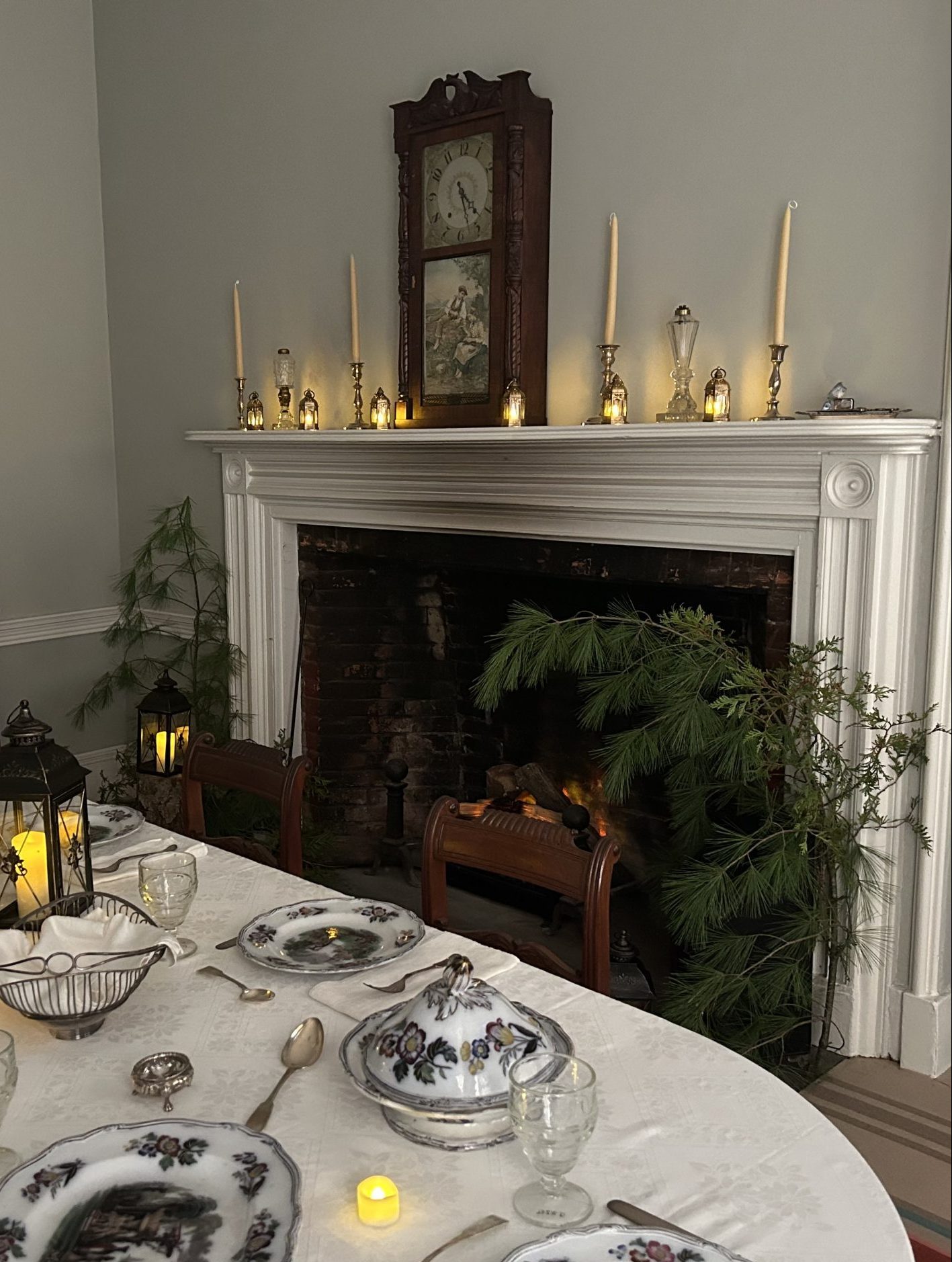 Image of a table set with fancy dish ware over a white tablecloth. Behind the table is a fireplace and mantel. On the mantel are candles and a rectangular wooden clock. On either side of the fireplace is a very small pine tree.